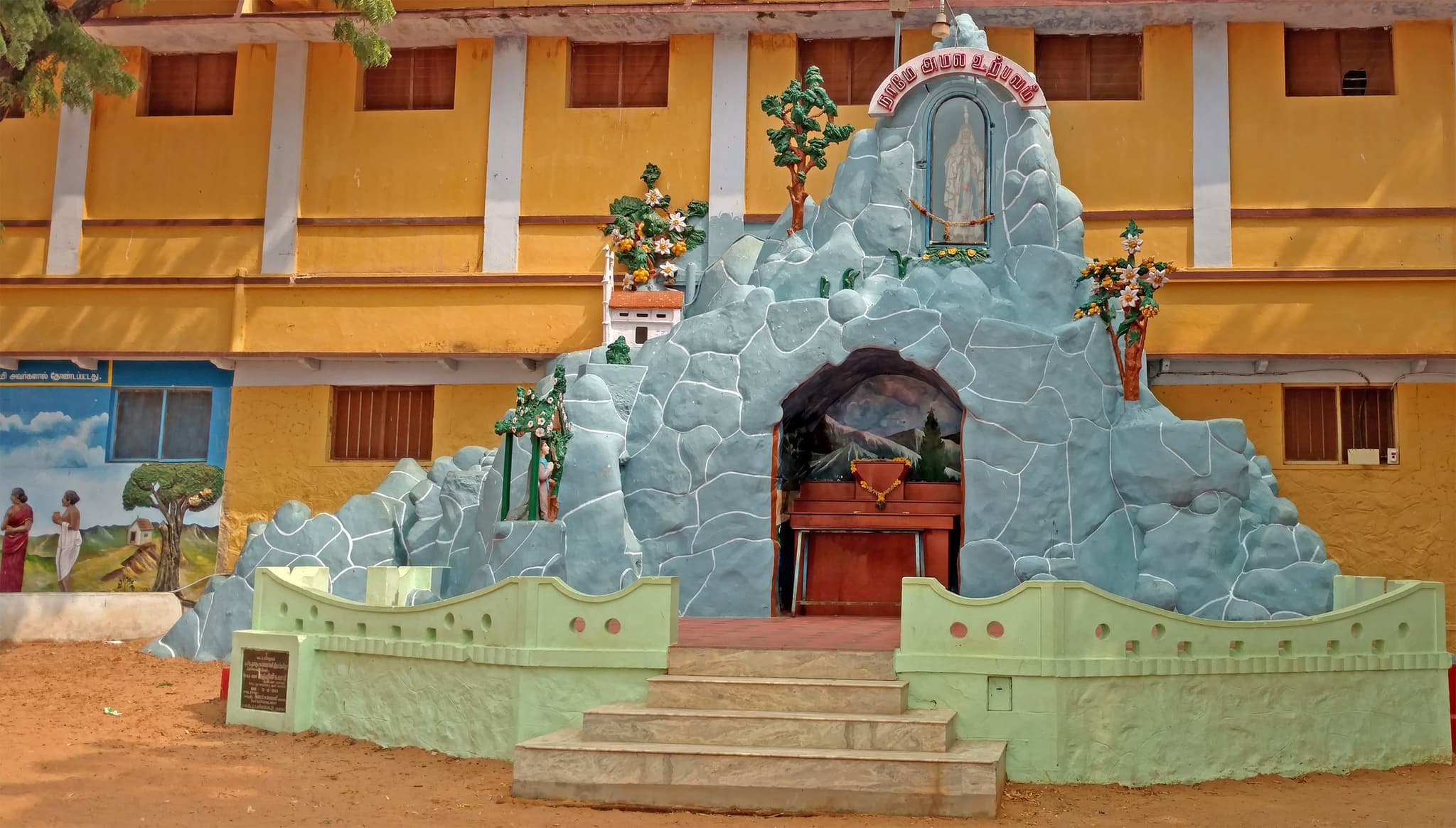 பங்கின் லூர்து குகை — நீண்ட மஞ்சள் நிறக் கட்டிடத்தின் முன் எழுப்பப்பட்ட வெளிர் நீலப் பாறைக் குகை, மேலுள்ள மாடத்திற்குப் படிகள் ஏறுகின்றன