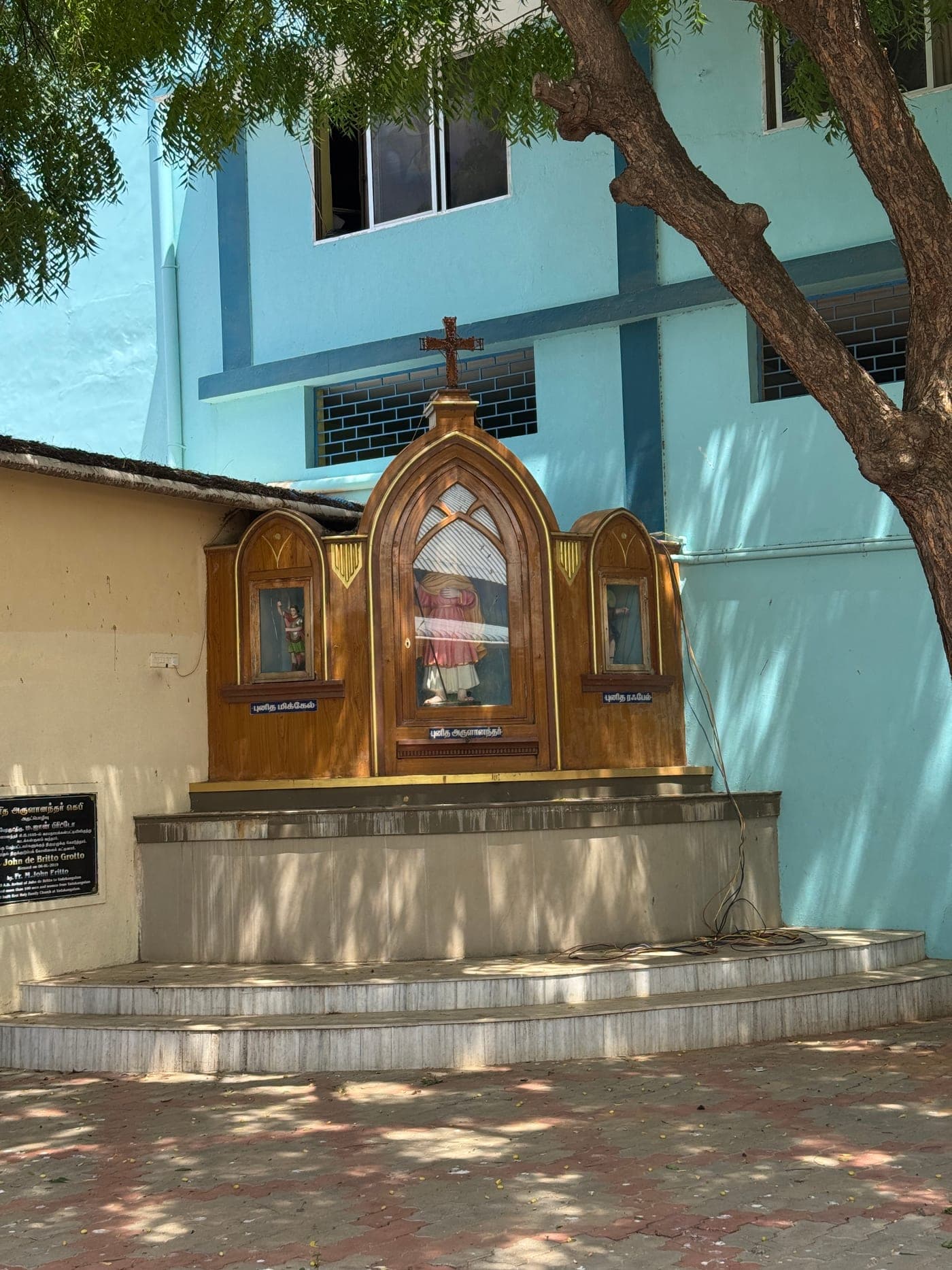 The John de Britto grotto in the grounds of the Holy Family Shrine, Vadakkankulam — a wooden Gothic shrine holding a statue of St John de Britto (Arulanandar), flanked by St Michael and St Raphael, beneath a neem tree