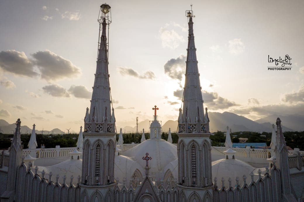 The twin spires and domed roof of the Holy Family Shrine at sunset, with the hills of the Western Ghats on the horizon